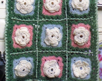 May include: A crocheted baby blanket with a green, pink, and blue color scheme. The blanket is made up of squares with white crocheted flowers in the center of each square.