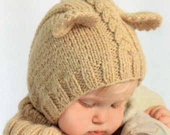 May include: A tan knitted baby hat with ear flaps and a matching neck warmer. The hat has a cable knit design on the crown. The baby is wearing a white shirt and holding a stuffed animal.