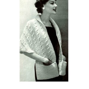 May include: A black and white image of a woman wearing a cream-coloured knitted shawl with pockets. The shawl has a textured cable knit design and a collar. The woman is wearing a black dress and has her hair styled up.