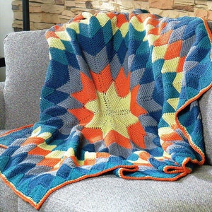 May include: A colorful crocheted throw blanket with a starburst pattern in shades of blue, orange, yellow, and gray. The blanket is folded and draped over a gray couch.