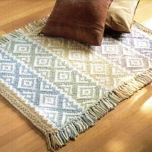 May include: A woven rug with a geometric pattern in shades of blue, green, and beige. The rug has a fringe border and is made of natural fibers.