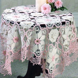 Könnte beinhalten: Ein runder, cremefarbener gehäkelter Tischläufer mit rosa gewelltem Rand und floralen Akzenten. Der Tischläufer ist über einem kleinen, schwarzen Tisch drapiert. Zwei Kerzen und rosa Blumen stehen darauf.