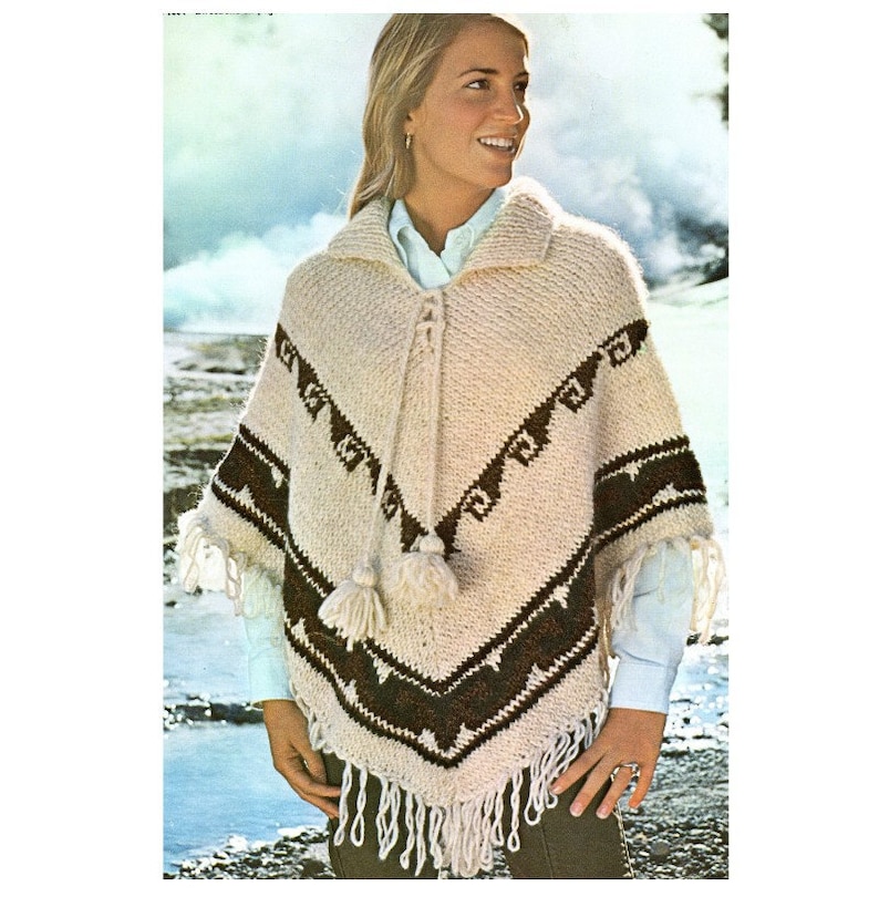 Op de afbeelding: Een cr&egrave;mekleurige gebreide poncho met een donkerbruin geometrisch patroon op de borst en de zoom. Het kledingstuk heeft een kraag, een vetersluiting aan de voorzijde met twee hangende kwastjes en lange franjes langs de onderrand.