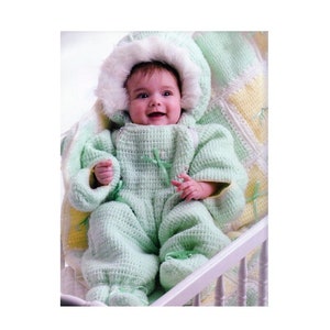 May include: A baby wearing a green crocheted jumpsuit with a white fur-trimmed hood. The baby is sitting in a white crib with a yellow and green crocheted blanket.