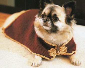May include: A small dog wearing a burgundy cape with gold trim and a decorative gold clasp. The dog has tan and black fur and is lying on a light-colored surface.
