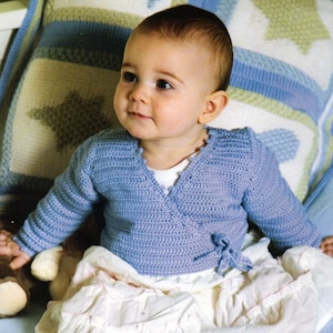 May include: A baby wearing a light blue crocheted cardigan with a white undershirt and a white skirt. The baby is sitting on a blue and white blanket with a stuffed animal.
