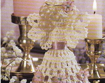May include: A white crocheted doll ornament with a flared skirt and a satin ribbon waist, positioned between two brass candle holders, each containing a lit pillar candle with a warm glow, creating a soft and illuminated decorative display.