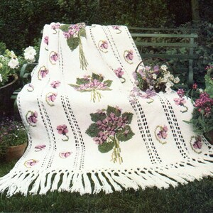 Pode incluir: Cobertor de crochê branco com um design floral com violetas roxas e verdes. O cobertor tem uma borda de franjas e é exibido em um banco de madeira.
