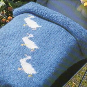 Pode incluir: Um cobertor de bebé azul de malha com um desenho de pato branco. O cobertor está dobrado ao meio e tem uma borda branca.