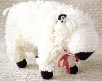 May include: A handmade white sheep plush toy with a fluffy, textured body. The sheep has black hooves, a black eye, and a red ribbon tied around its neck. The toy is made of soft, white yarn.