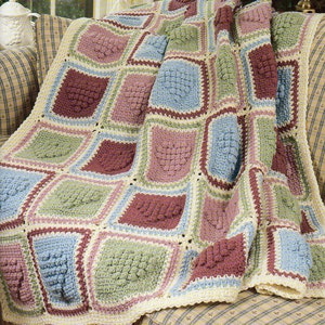 May include: A handmade crochet blanket in a patchwork design. The blanket features squares in shades of blue, green, pink, and burgundy, bordered by cream and light pink. The blanket is draped over a plaid armchair.