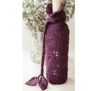 Puede incluir: Una funda de botella de vino de punto, de color púrpura oscuro, con un diseño decorativo de hojas. La funda tiene una larga correa trenzada y adornos en forma de hoja. La botella es parcialmente visible en la parte superior.