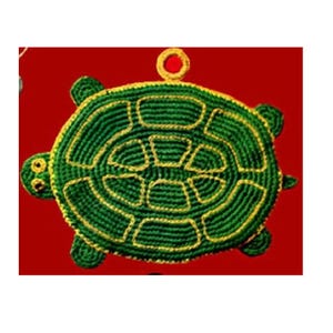 Op de afbeelding: Een groen en geel gehaakt schildpadornament met een lus om op te hangen. De schildpad heeft een gedetailleerd schildpatroon en kleine, ronde ogen. De achtergrond is effen rood.