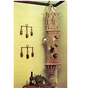May include: A beige macrame wine rack, designed to hold several bottles, hangs on a wall. The rack has diamond patterns and is made of rope. Two sets of bronze bells are mounted on the wall. A bottle and glass are on a table below.