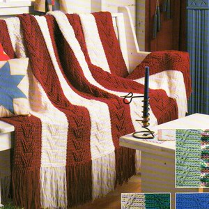 May include: A red and white crocheted throw blanket with fringe, draped over a white sofa. The blanket has a cable knit pattern and is reminiscent of the American flag.