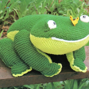May include: A large, handmade green frog plush toy with yellow accents on its belly and feet. The frog has large white eyes, a smiling mouth, and a small yellow butterfly on its head. The toy is made of knitted fabric and sits on a wooden surface.