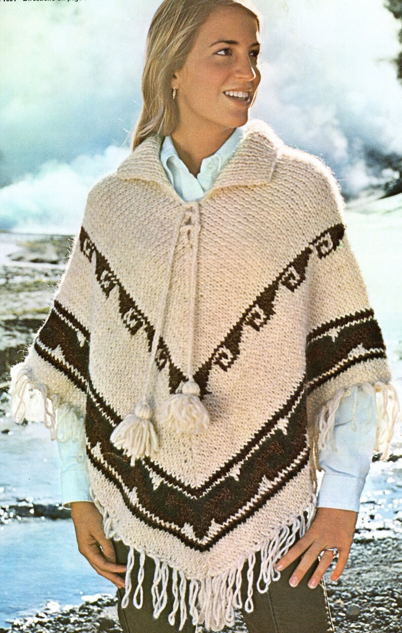 Op de afbeelding: Een cr&egrave;mekleurige gebreide poncho met een donkerbruin geometrisch patroon, een kraag en een vetersluiting aan de voorzijde met twee bijpassende cr&egrave;mekleurige kwastjes. De zoom is afgewerkt met lange, dikke franjes aan de onderkant.