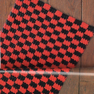 Op de afbeelding: Een rood en zwart geblokt tapijt met een herhalend patroon. Het tapijt is rechthoekig en lijkt gemaakt te zijn van een geweven materiaal. Het tapijt ligt op een houten oppervlak.