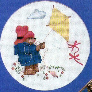 May include: Cross-stitch design featuring two teddy bears in blue coats and pink hats, flying a yellow kite. The bears stand in a field with flowers and grass, with a small cloud in the sky. The design is set against a white background.