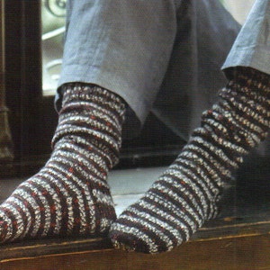 Puede incluir: Primer plano de dos pies con calcetines a rayas. Los calcetines son principalmente de color marrón oscuro con rayas horizontales blancas y rojas. La persona lleva pantalones grises. Los calcetines parecen estar tejidos o ganchillados.