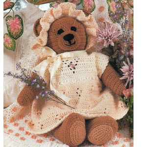 May include: A handmade crochet teddy bear wearing a cream-colored dress and bonnet. The bear has brown fur, a black stitched nose and eyes, and is holding a small bouquet of purple flowers. The bear is sitting on a floral patterned surface.