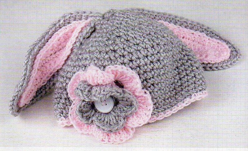 Pode incluir: Um chap&eacute;u de coelho de croch&ecirc; feito &agrave; m&atilde;o, cinza com detalhes em rosa. O chap&eacute;u apresenta orelhas longas e verticais com forro interno rosa e uma flor decorativa com um bot&atilde;o no centro. Para beb&ecirc;s e crian&ccedil;as pequenas.