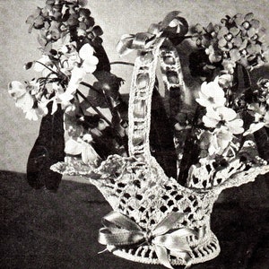 May include: A crocheted basket with a handle, filled with flowers. The basket has a decorative openwork design and is adorned with a satin ribbon bow. The flowers are a mix of white and purple.