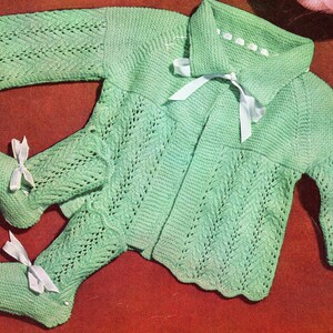 May include: A mint green knitted baby outfit, including a jacket with a collar and a pair of booties. The outfit features a delicate openwork design and is adorned with white ribbon bows. The outfit is a vintage design.