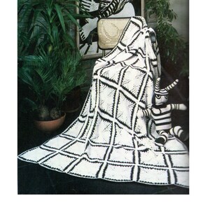 Puede incluir: Una manta de crochet en blanco y negro con un patrón de diamantes. La manta está drapeada sobre una silla y un juguete a rayas está sentado sobre la manta.