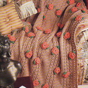 May include: A crocheted blanket in a light brown hue, adorned with numerous red rose appliqués. The blanket is draped over a chair, with a matching pillow and a small fringed bag. An open book rests nearby.