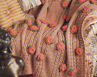 May include: A crocheted blanket in a light brown hue, adorned with numerous red rose appliqués. The blanket is draped over a chair, with a matching pillow and a small fringed bag. An open book rests nearby.