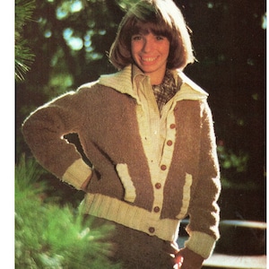 Puede incluir: Un cárdigan de punto marrón y crema con cuello, cierre de botones y bolsillos. El cárdigan tiene ribetes color crema en el cuello, los bolsillos y los puños. La persona lleva una camisa a cuadros debajo.