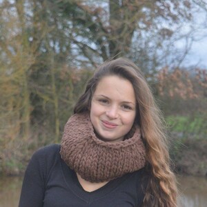 Puede incluir: Una persona joven con cabello largo y castaño que lleva una bufanda de punto infinito marrón y una camisa negra. La persona está de pie frente a un fondo borroso de árboles y agua.