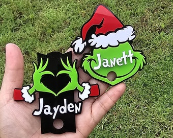 May include: Two Christmas-themed ornaments are held in a hand. One is a green Grinch-like character with the name "Janett" in white. The other is a black ornament with a green heart and the name "Jayden". Both have a hole.