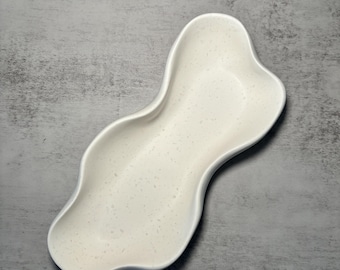 May include: White ceramic serving dish with a wavy, organic shape. The dish is designed to hold small items like snacks or jewelry.