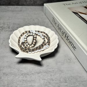 May include: Two beaded bracelets with white and gray beads, a white stone charm, and silver accents, displayed on a white ceramic seashell dish.