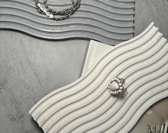May include: Two wavy, gray and white ceramic trays with raised lines. The gray tray has a silver beaded necklace with white letter beads spelling "WISH" on it. The white tray has a ring with a white heart-shaped stone on it.