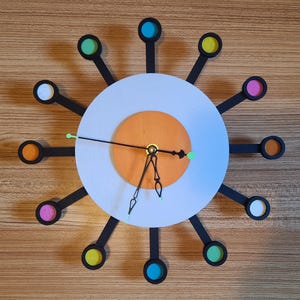 May include: A white and orange clock with black arms and a black frame. The frame has 12 colorful circles around the clock face.