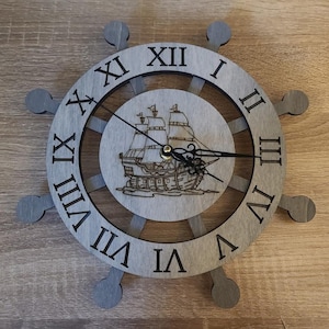 May include: A gray wooden clock shaped like a ship's wheel with Roman numeral markings and a ship design in the center.