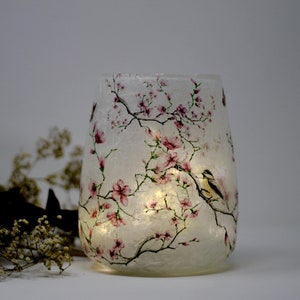 May include: A white glass candle holder with a floral design featuring pink flowers and birds. The candle holder is illuminated from within, creating a warm glow.