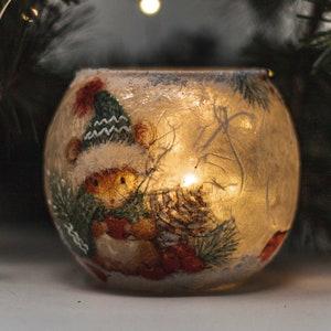 Peut inclure: Un photophore rond en verre avec un motif festif. Le photophore présente une scène hivernale avec un ours portant un chapeau et une écharpe. La bougie à l'intérieur diffuse une lueur chaleureuse, entourée de branches de conifères.