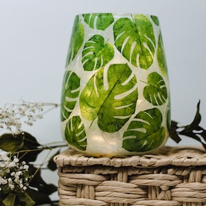 Può includere: Un portacandele in vetro trasparente con sfondo bianco e design a foglie tropicali verdi. Il portacandele è appoggiato su una scatola di vimini intrecciata.