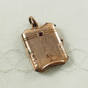 Peut inclure: Un médaillon en or avec un design Art Déco et une pierre précieuse rouge. Le médaillon a un couvercle à charnière et une boucle pour attacher une chaîne.