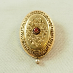 Puede incluir: Un medallón ovalado de oro con un diseño intrincado y una piedra preciosa roja en el centro. El medallón tiene un colgante de perla.