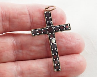 May include: A cross pendant featuring a series of dark red, round-cut garnet stones set in a metallic frame. The cross has a small circular jump ring at the top for attachment. The stones are arranged in a single row along the arms of the cross.
