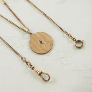Könnte beinhalten: Eine goldene Kette mit einem runden Anhänger, der mit einem kleinen roten Stein verziert ist. Die Kette hat einen Verschluss und einen kleinen Haken am Ende.