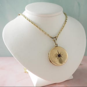 Puede incluir: Un colgante de medallón circular en tono dorado suspendido de una cadena de eslabones a juego. La parte frontal del medallón presenta un diseño de estrella de ocho puntas grabado en el centro con una pequeña piedra oscura incrustada.