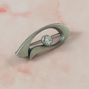 Puede incluir: Broche plateado con un diseño curvo y abierto. Una gema redonda y transparente está engastada en el centro. El broche tiene un borde texturizado y un cierre de alfiler. Una joya clásica.