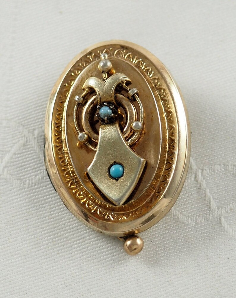 K&ouml;nnte beinhalten: Ovales goldenes Medaillon mit einem detaillierten Design mit einer T&uuml;rkis- und Perlenverzierung.