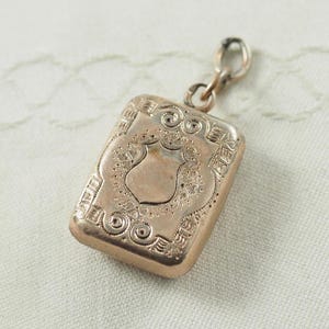 Könnte beinhalten: Ein goldfarbener Anhänger mit einem verzierten Design, das ein Schild und eine Kette zeigt. Der Anhänger ist rechteckig und hat ein detailliertes, graviertes Muster.